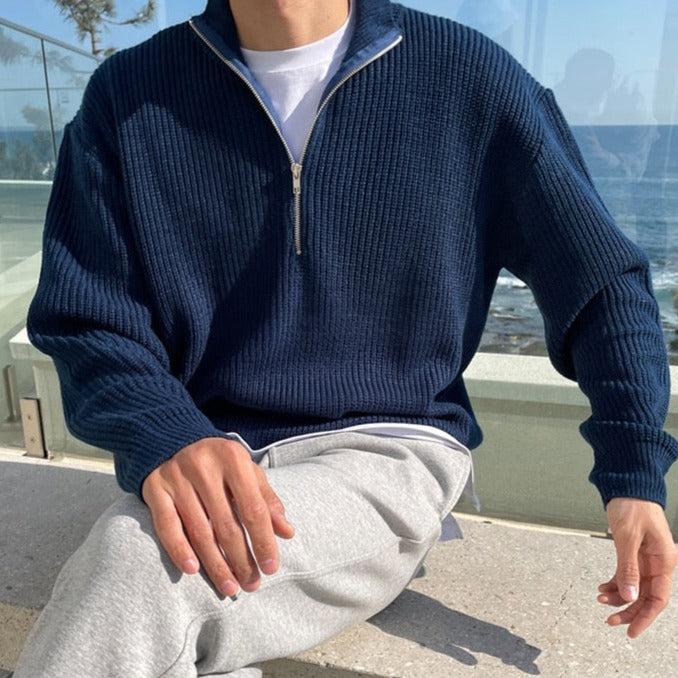 Uomo con maglione blu a collo alto con zip, seduto all'aperto. Indossa pantaloni grigi. Sfondo di mare e cielo. Moda casual maschile.