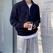 Uomo con maglione blu scuro a collo alto e zip, pantaloni grigi, tiene una tazza da caffè. Look casual autunnale. Sfondo urbano minimalista.