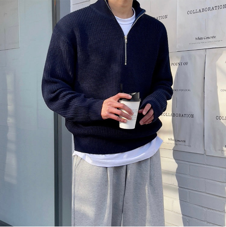 Uomo con maglione blu scuro a collo alto e zip, pantaloni grigi, tiene una tazza da caffè. Look casual autunnale. Sfondo urbano minimalista.