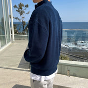 Uomo con maglione blu scuro in lana, vista mare. Moda maschile autunnale, abbigliamento casual elegante, sfondo costiero.