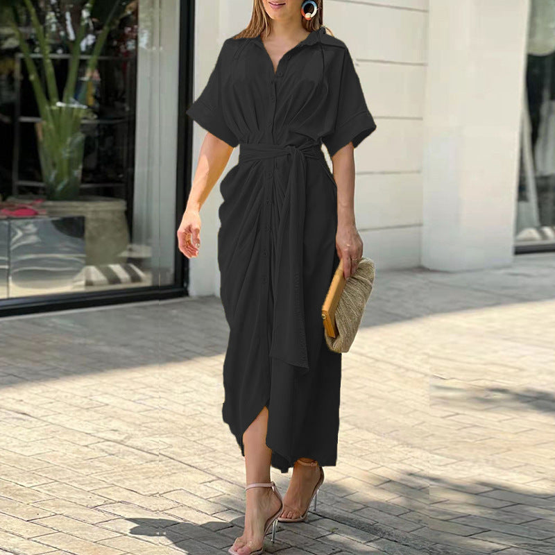 Donna con abito lungo nero elegante, cintura in vita, sandali con tacco, borsa a mano beige, passeggia all'aperto. Moda estiva femminile chic.