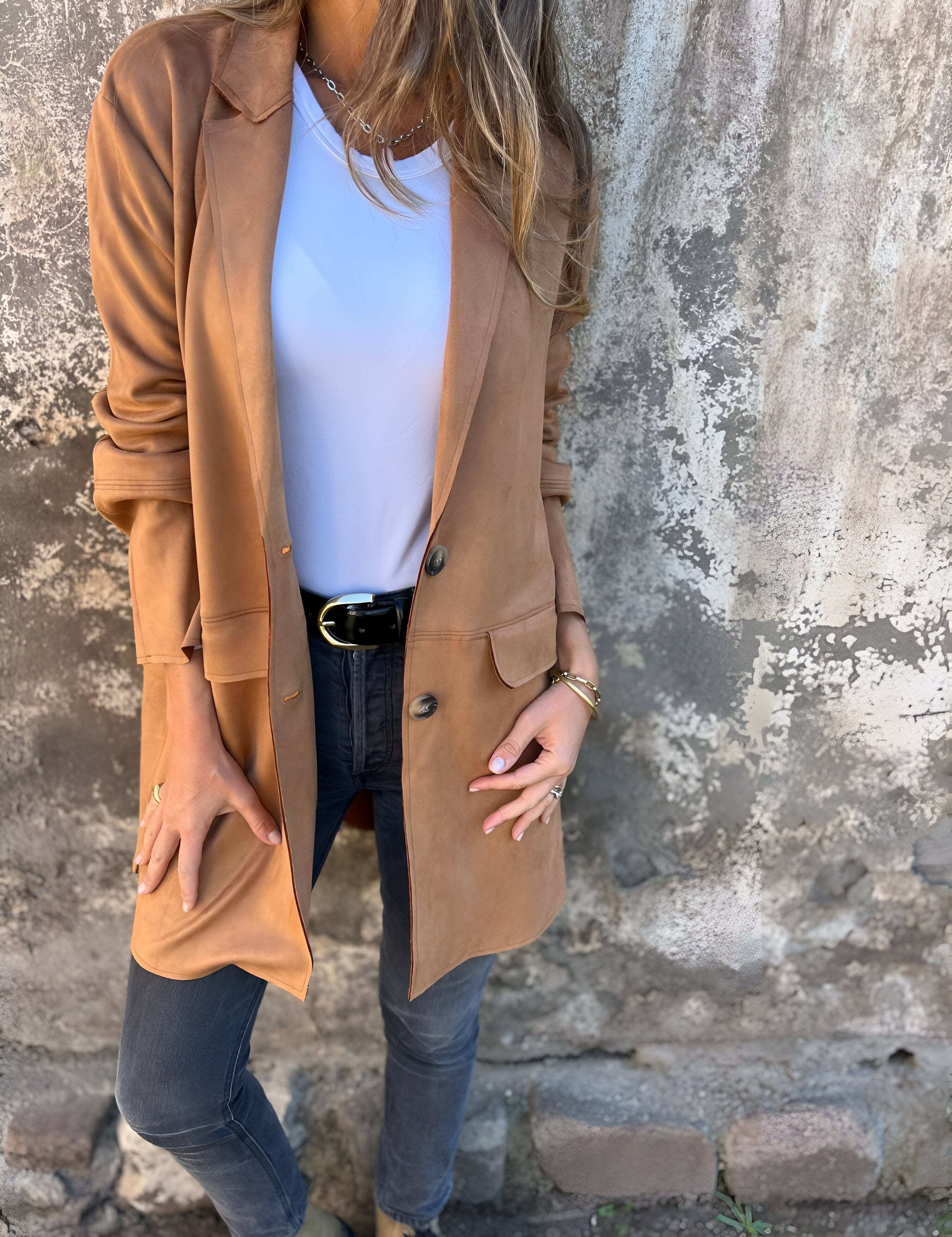 Donna con giacca lunga marrone, maglietta bianca e jeans neri, posa contro un muro rustico. Moda autunnale casual, stile urbano chic.