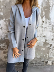 Donna indossa cappotto grigio con bottoni, su sfondo di muro rustico. Moda autunnale, abbigliamento casual elegante, stile urbano.