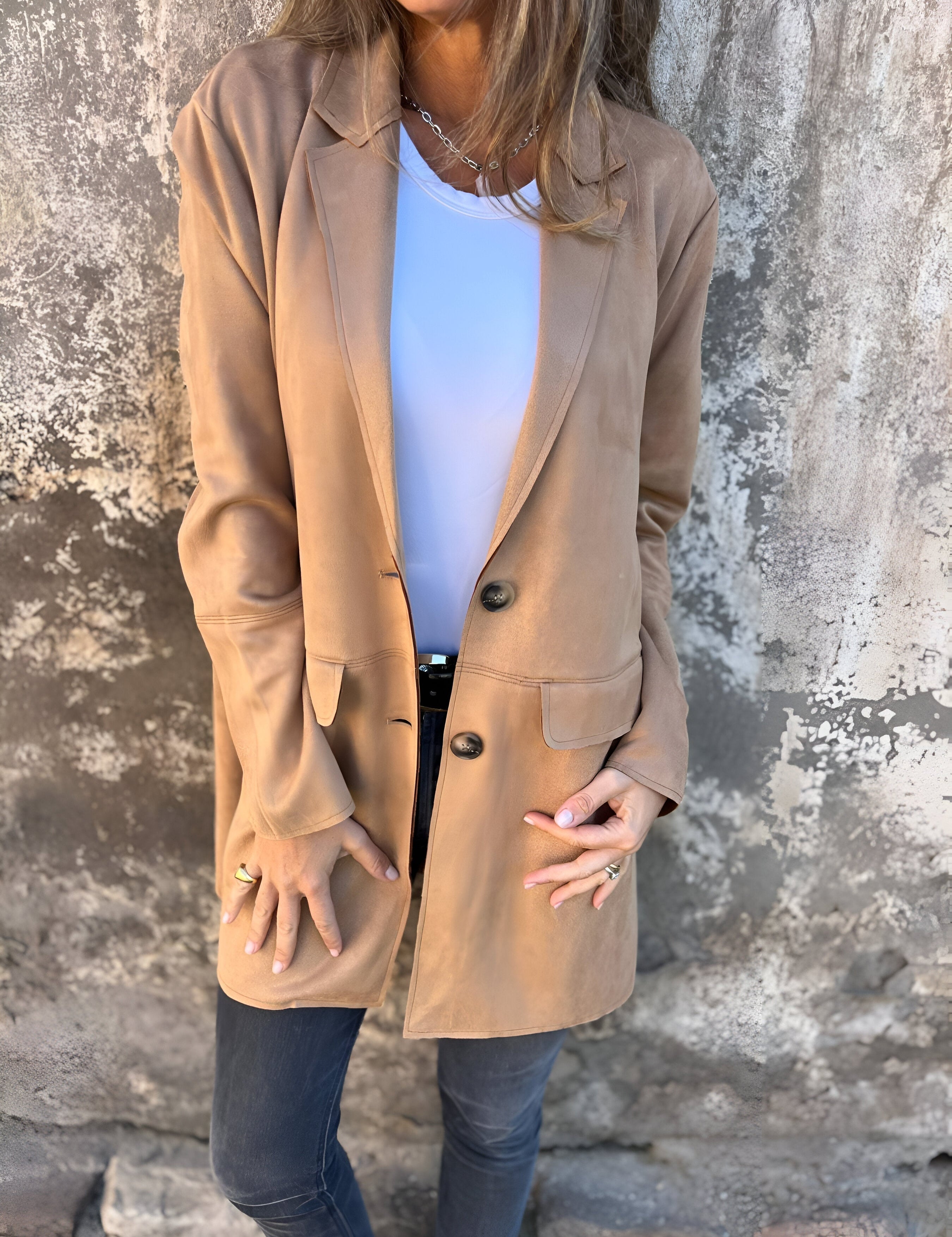 Donna con cappotto beige elegante, camicia bianca e jeans scuri, posa contro un muro rustico. Moda autunnale, stile casual chic.