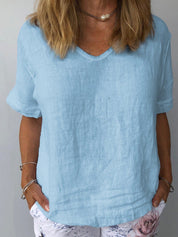 Donna con maglietta di lino azzurra, scollo a V, maniche corte, abbinata a pantaloni bianchi con stampa floreale. Moda estiva casual elegante.