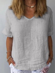Donna con maglietta di lino grigia, scollo a V, maniche corte, indossa braccialetti e collana, pantaloni bianchi con stampa floreale.