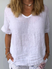 Donna con maglietta bianca in lino, scollo a V, maniche corte. Stile casual estivo, abbinata a pantaloni chiari. Accessori: bracciali e collana.