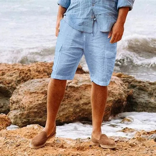 Uomo in pantaloncini di lino azzurri e camicia abbinata su spiaggia rocciosa, indossa mocassini marroni. Look estivo casual maschile.