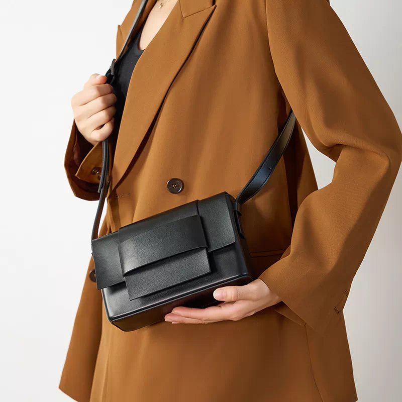 Donna con cappotto marrone indossa borsa a tracolla nera in pelle, design moderno e minimalista. Accessorio moda elegante per look autunnali.