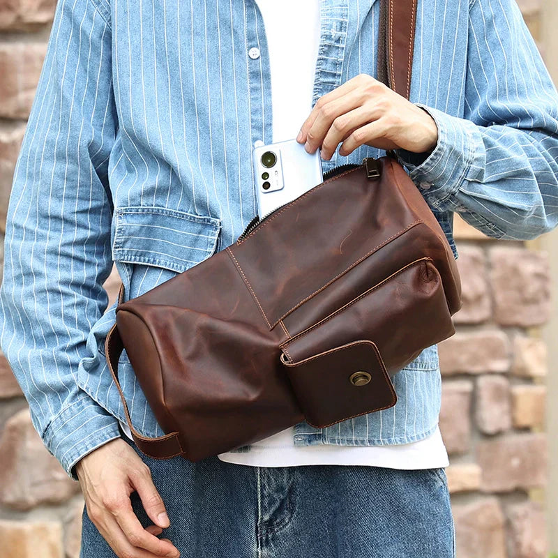 Uomo con giacca di jeans tiene borsa a tracolla in pelle marrone, tasca frontale con bottone, inserisce smartphone. Accessorio moda casual.