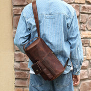 Uomo con giacca di jeans indossa borsa a tracolla in pelle marrone, design vintage, tasca frontale con bottone, sfondo muro di mattoni.