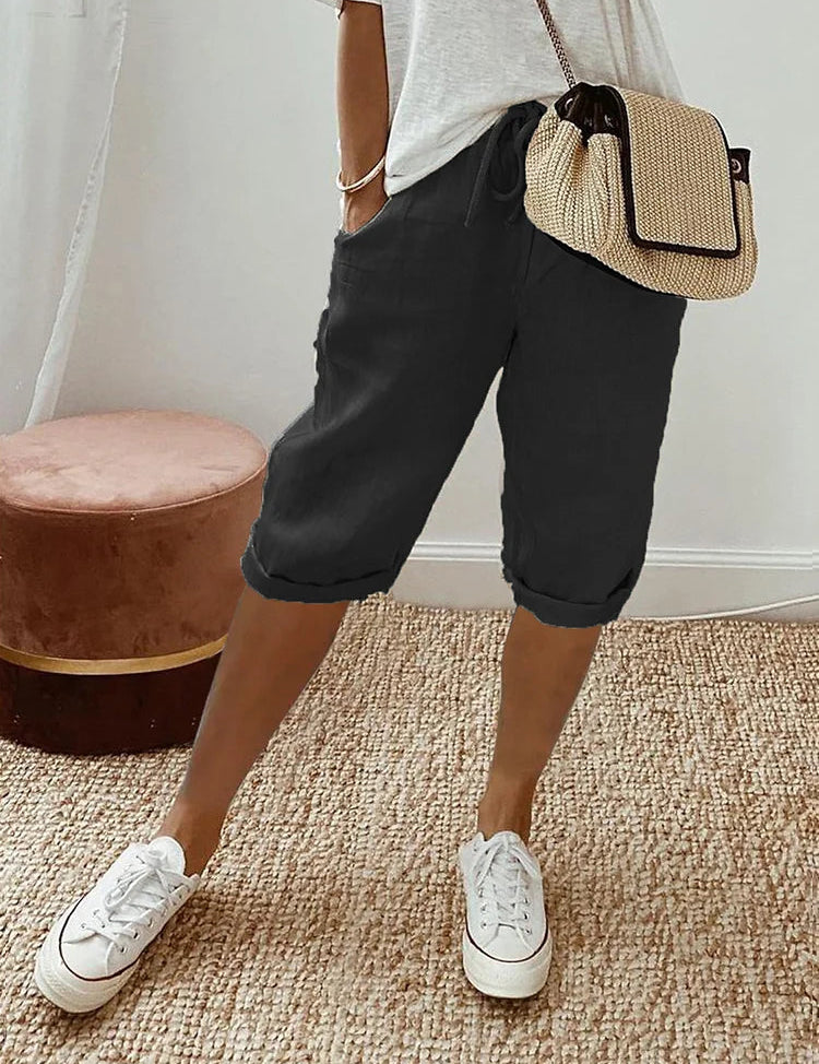 Pantaloncini neri casual da donna, abbinati a maglietta bianca e borsa a tracolla beige, su tappeto intrecciato. Scarpe da ginnastica bianche.