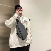 Uomo con felpa bianca oversize, cappello nero e borsa a tracolla grigia, scatta selfie con smartphone rosso. Stile casual moderno.