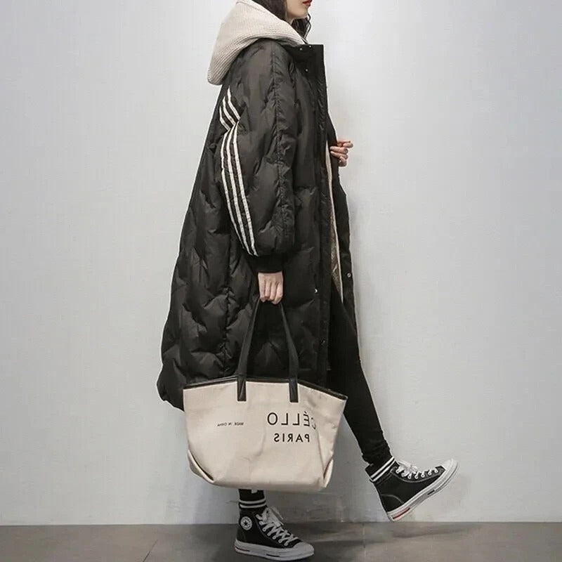 Cappotto lungo nero trapuntato con cappuccio, dettagli a righe bianche, abbinato a borsa beige e sneakers nere. Moda invernale casual donna.