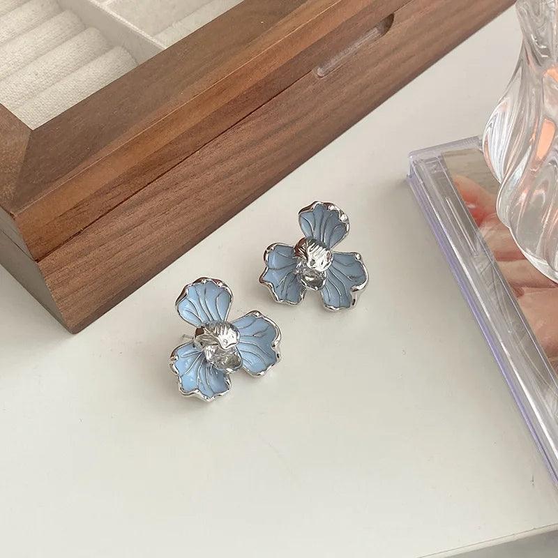 Orecchini a forma di fiore blu con dettagli argentati su sfondo chiaro, accanto a una scatola di legno. Accessori moda eleganti e raffinati.