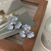 Orecchini a forma di fiore blu su sfondo di libri e candela. Gioielli eleganti, design floreale, accessori moda donna.