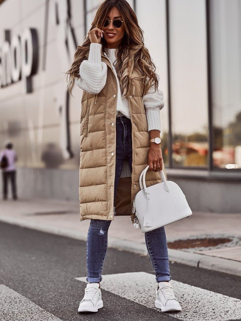 Donna con cappotto lungo beige, maglione bianco, jeans attillati e sneakers bianche, tiene una borsa bianca. Moda autunnale urbana.