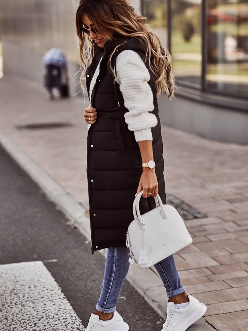 Donna con gilet lungo nero imbottito, maglione bianco, jeans aderenti e sneakers bianche, borsa bianca. Moda autunnale casual chic.