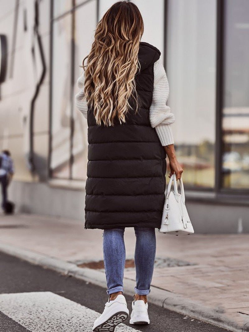Donna di spalle con gilet lungo nero imbottito, jeans aderenti, sneakers bianche e borsa bianca. Moda autunnale casual in città.