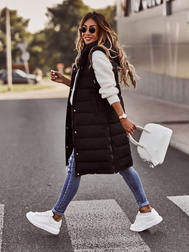 Donna alla moda con gilet lungo nero, jeans attillati e sneakers bianche, attraversa la strada con borsa bianca. Look casual autunnale.