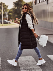 Donna alla moda con gilet lungo nero, jeans attillati e sneakers bianche, attraversa la strada con borsa bianca. Look casual autunnale.