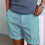 Uomo con pantaloncini azzurri in lino, maglietta grigia, mani in tasca. Moda estiva casual, abbigliamento maschile elegante.