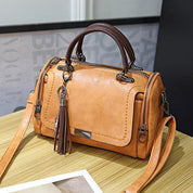 Borsa a mano in pelle marrone chiaro con manici scuri, cerniere laterali e nappina decorativa, su tavolo di legno. Accessorio moda elegante.