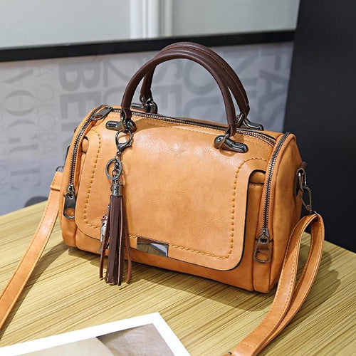 Borsa a mano in pelle marrone chiaro con manici scuri, cerniere laterali e nappina decorativa, su tavolo di legno. Accessorio moda elegante.
