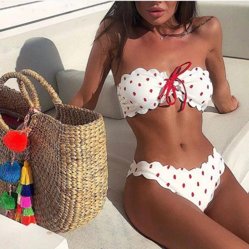 Donna in bikini bianco a pois rossi con fiocco rosso, seduta su divano esterno. Borsa di paglia con pompon colorati a lato. Moda mare estiva.