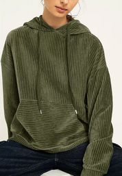 Felpa verde oliva a coste con cappuccio e tasca frontale, indossata da una persona seduta. Stile casual, tessuto morbido e confortevole.