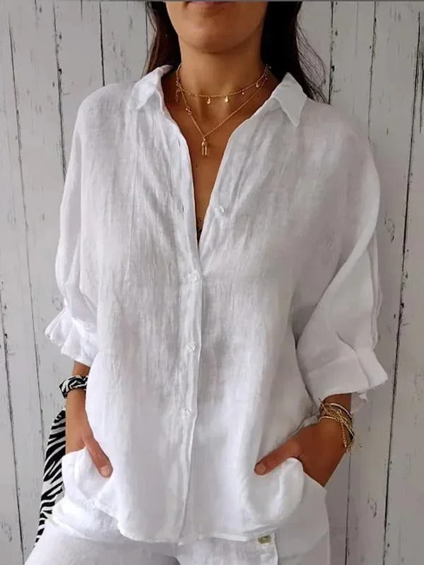 Camicia bianca in lino da donna, maniche lunghe, stile casual, abbinata a collane dorate. Sfondo in legno chiaro. Moda estiva elegante.