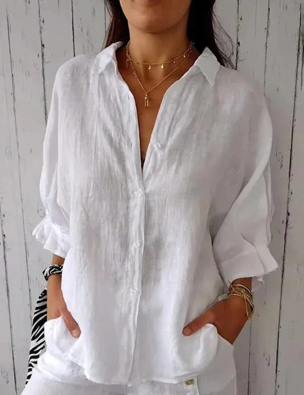 Camicia bianca in lino da donna, maniche lunghe, stile casual, abbinata a collane dorate. Sfondo in legno chiaro. Moda estiva elegante.