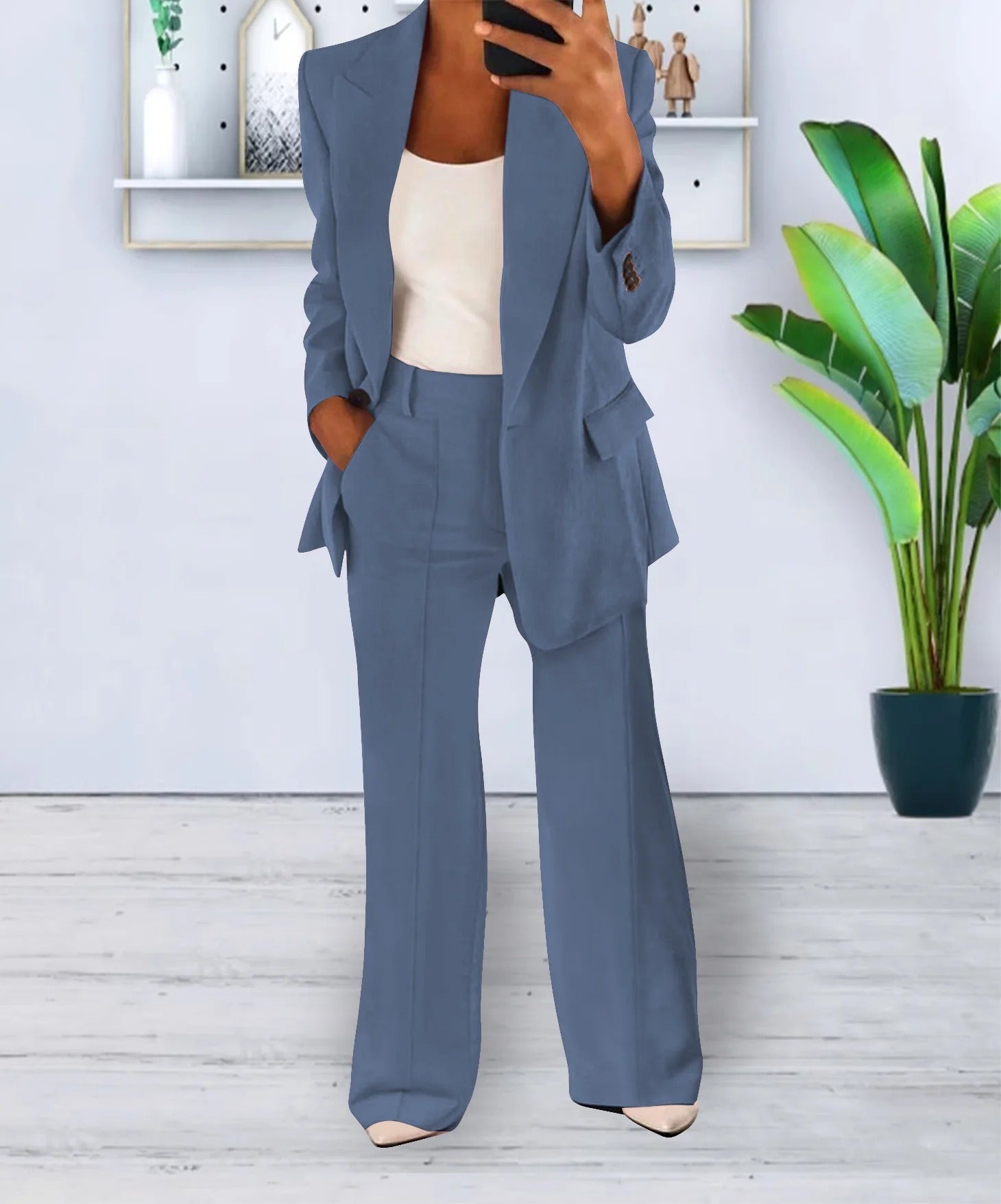 Completo donna elegante blu, giacca e pantaloni a zampa, indossato con top bianco. Sfondo moderno con pianta verde e scaffale decorativo.