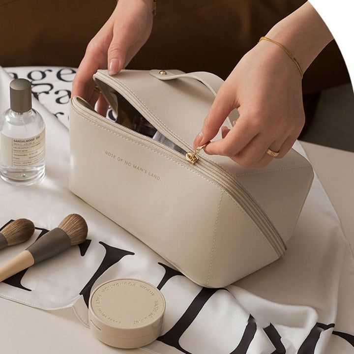 Borsa cosmetica beige elegante con zip, circondata da pennelli trucco, flacone profumo e crema. Accessori bellezza essenziali per viaggio.