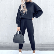 Donna con tuta nera casual, borsa a mano nera, sfondo grigio. Moda autunnale, abbigliamento comodo, stile urbano.